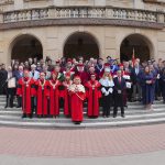 Grupowe zdjęcie uczestników uroczystości akademickiej wykonane przed głównym wejściem do budynku Uniwersytetu Jana Kochanowskiego w Kielcach. Na schodach stoją osoby w togach i strojach ceremonialnych oraz zaproszeni goście.