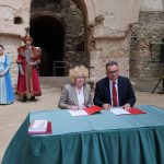 Dwie osoby siedzą przy stole nakrytym turkusowym obrusem i podpisują dokumenty w zabytkowym, częściowo zrujnowanym wnętrzu z kamiennymi ścianami oraz arkadami. Po lewej stronie stoi osoba w ciemnym garniturze, a w tle widoczne są dwie osoby w historycznych strojach. Obok stołu leżą czerwone teczki z dokumentami.