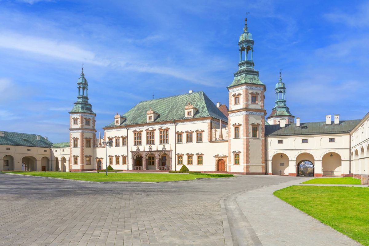 Jasny, barokowy pałac z dwiema wieżami z zielonymi hełmami po bokach. Elewacja z czerwoną cegłą, centralny portyk z arkadami. Przed budynkiem dziedziniec z kostki brukowej, trawniki i błękitne niebo z chmurami. Po bokach skrzydła z podcieniami.