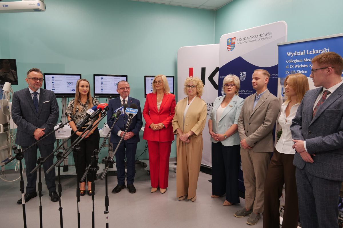 Uczestnicy konferencji prasowej w sali dydaktycznej z ekranami medycznymi. Dziewięć osób stoi w rzędzie przed mikrofonami. W tle ścianka z logo Urzędu Marszałkowskiego Województwa Świętokrzyskiego oraz banner Wydziału Lekarskiego UJK.