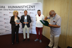 Cztery osoby na tle ścianki Wydziału Humanistycznego UJK w Kielcach; dwoje z nich trzyma w rękach publikacje, a mężczyzna po prawej przegląda zieloną teczkę.