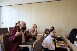 Uczestnicy spotkania w sali wykładowej; na pierwszym planie mężczyzna w bordowej marynarce i okularach gestykuluje podczas wypowiedzi.