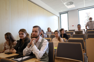 Młodzi ludzie i studenci siedzący w ławkach nowoczesnej sali wykładowej, słuchający prelekcji.