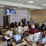 Widok od tyłu sali wykładowej na zgromadzoną publiczność oraz prelegentów i ekran multimedialny z przodu auli.