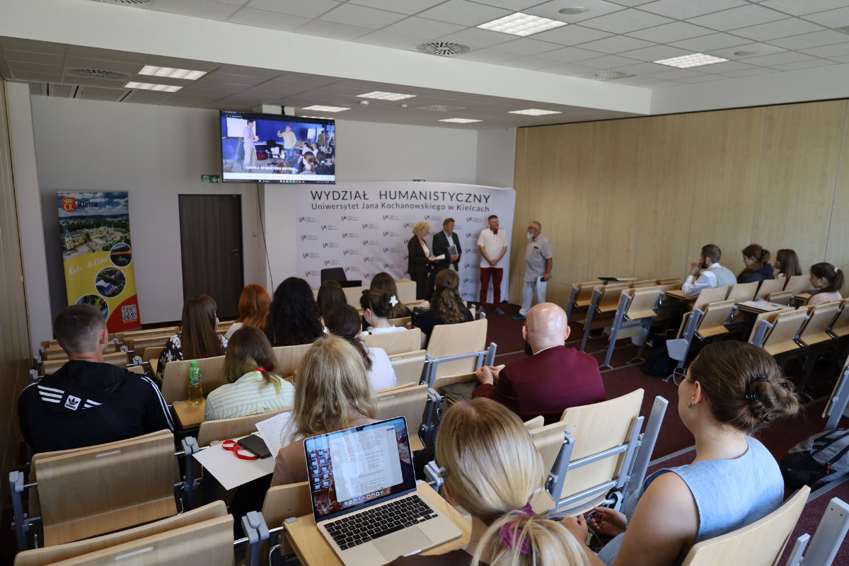 Widok od tyłu sali wykładowej na zgromadzoną publiczność oraz prelegentów i ekran multimedialny z przodu auli.