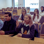 Zróżnicowana grupa studentów siedzi w przednich rzędach sali wykładowej. Studentka z różowymi włosami podnosi rękę, zgłaszając się do zadania pytania. Pozostali studenci słuchają, w tym kobieta w jasnoniebieskim hidżabie i mężczyzna siedzący obok niej.