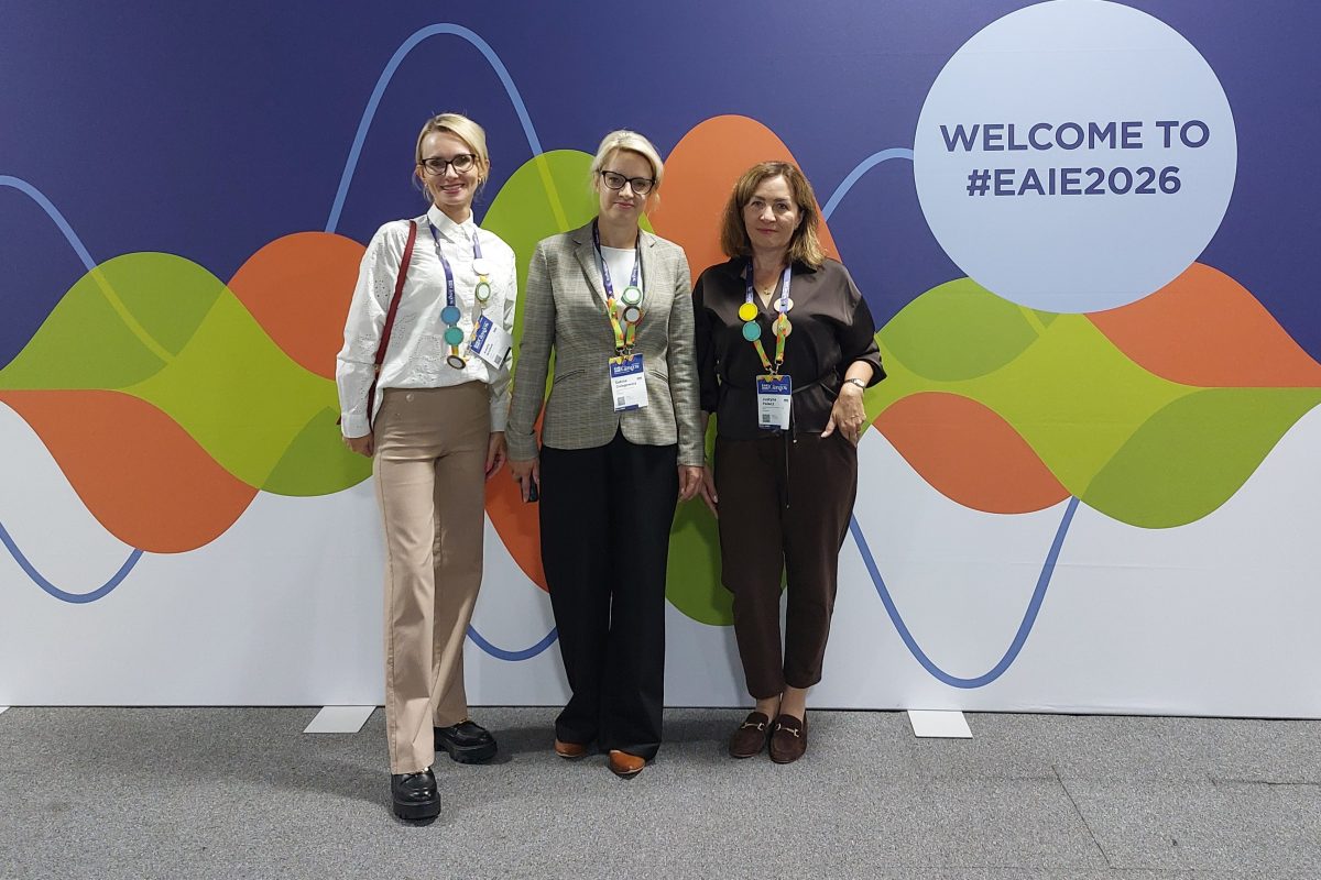 Trzy kobiety stoją uśmiechnięte na tle kolorowej ścianki konferencyjnej z napisem „WELCOME TO #EAIE2026”. Wszystkie mają na szyi identyfikatory na smyczach. Są ubrane w eleganckie, biznesowe stroje: koszule, żakiet oraz spodnie w odcieniach beżu, czerni i brązu.