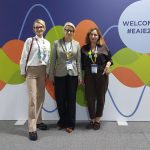 Trzy kobiety stoją uśmiechnięte na tle kolorowej ścianki konferencyjnej z napisem „WELCOME TO #EAIE2026”. Wszystkie mają na szyi identyfikatory na smyczach. Są ubrane w eleganckie, biznesowe stroje: koszule, żakiet oraz spodnie w odcieniach beżu, czerni i brązu.