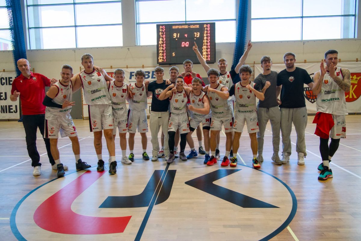 Radosna, młoda drużyna koszykarska (w białych strojach) i sztab świętują na parkiecie hali sportowej. Stoją w luźnej grupie, uśmiechają się, obejmują i wykonują gesty triumfu. W tle widać tablicę wyników z rezultatem 91:73, a na parkiecie przed nimi znajduje się duże logo "UJK" z czerwoną i czarną grafiką.