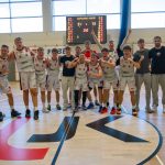 Radosna, młoda drużyna koszykarska (w białych strojach) i sztab świętują na parkiecie hali sportowej. Stoją w luźnej grupie, uśmiechają się, obejmują i wykonują gesty triumfu. W tle widać tablicę wyników z rezultatem 91:73, a na parkiecie przed nimi znajduje się duże logo "UJK" z czerwoną i czarną grafiką.