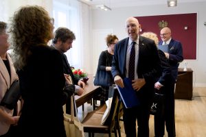 Stojący ambasador z niebieską teczką i torbą prezentową, uśmiechający się do grupy osób zgromadzonych w gabinecie.