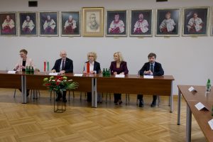 5 osób za stołem prezydialnym z flagami Włoch i Polski, w sali ozdobionej portretami rektorów.