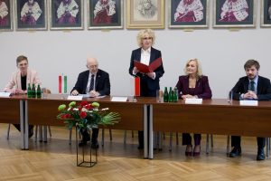 Rektor uczelni wygłasza przemówienie na stojąco, trzymając czerwoną teczkę, podczas gdy siedzący obok ambasador słucha.