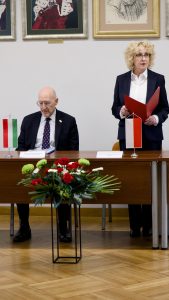 Rektor wygłaszająca przemówienie z czerwoną teczką w dłoni, stojąca obok siedzącego ambasadora za stołem z flagami Włoch i Polski.