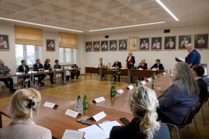 Szeroki kadr sali, na którym widać zgromadzonych gości i władze uczelni podczas oficjalnego spotkania z układem stołów w podkowę.