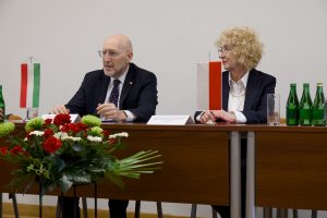 Przemawiający ambasador siedzący za stołem prezydialnym z kwiatami, w towarzystwie słuchającej go Rektor.