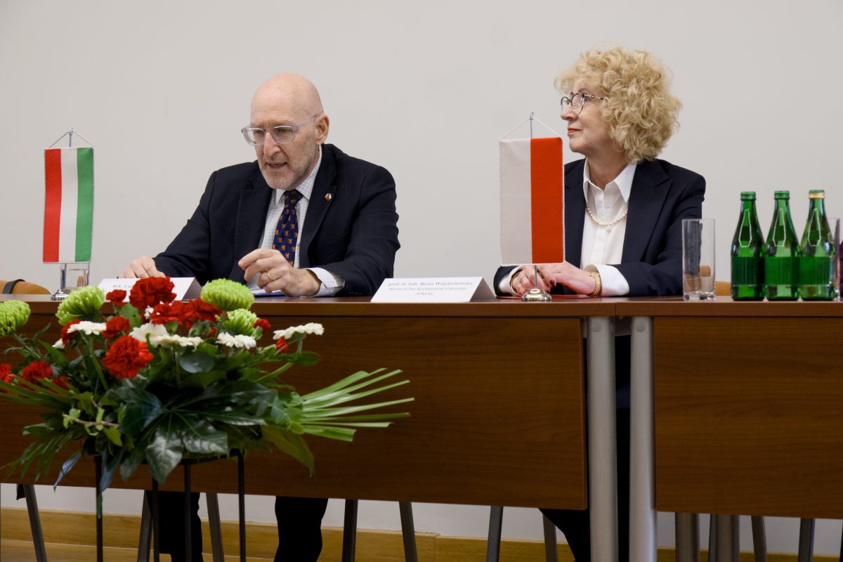 Przemawiający ambasador siedzący za stołem prezydialnym z kwiatami, w towarzystwie słuchającej go Rektor.
