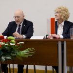 Przemawiający ambasador siedzący za stołem prezydialnym z kwiatami, w towarzystwie słuchającej go Rektor.