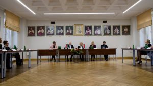 Szeroki kadr sali, na którym widać zgromadzonych gości i władze uczelni podczas oficjalnego spotkania z układem stołów w podkowę.