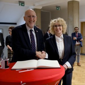 Uśmiechnięty ambasador wymieniający uścisk dłoni z Rektor tuż nad podpisaną księgą.
